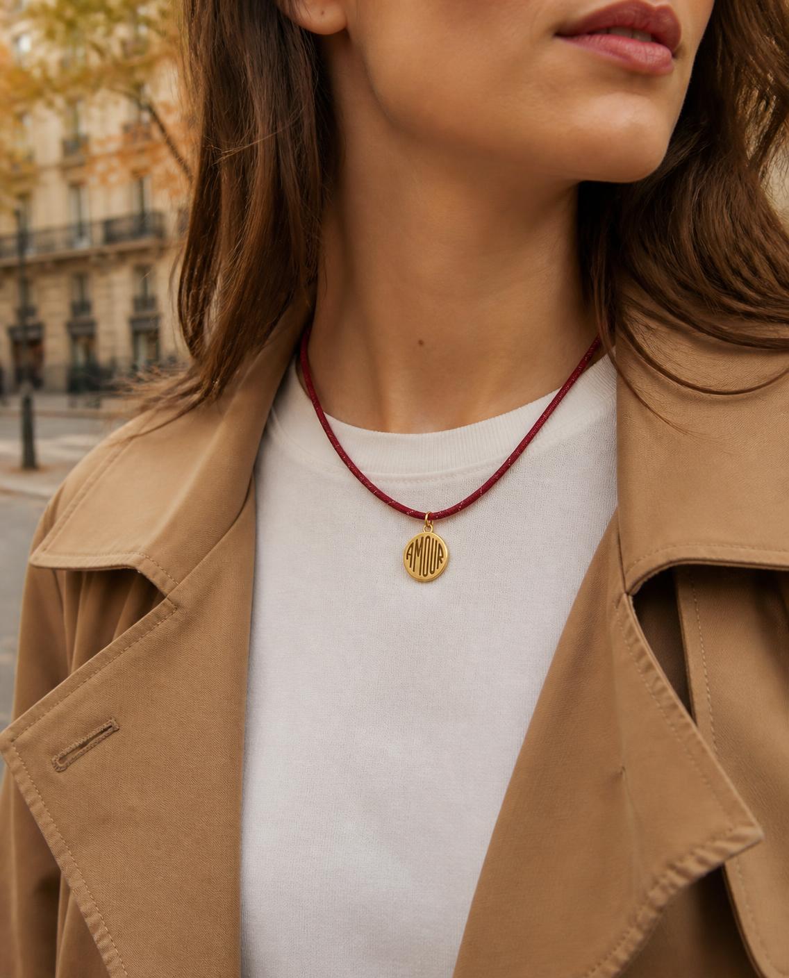 Woman wearing the Isla Sol Amour Cord Necklace, burgundy paracord with a gold AMOUR medallion, with a white tee and camel trench coat