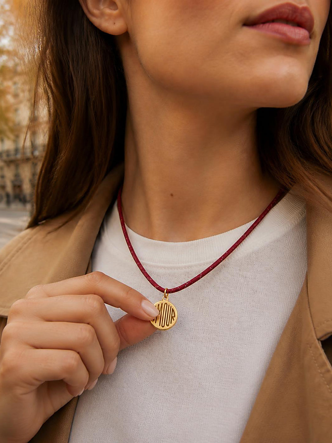 Close-up of a woman holding the gold AMOUR medallion on the Isla Sol Amour Cord Necklace, burgundy paracord