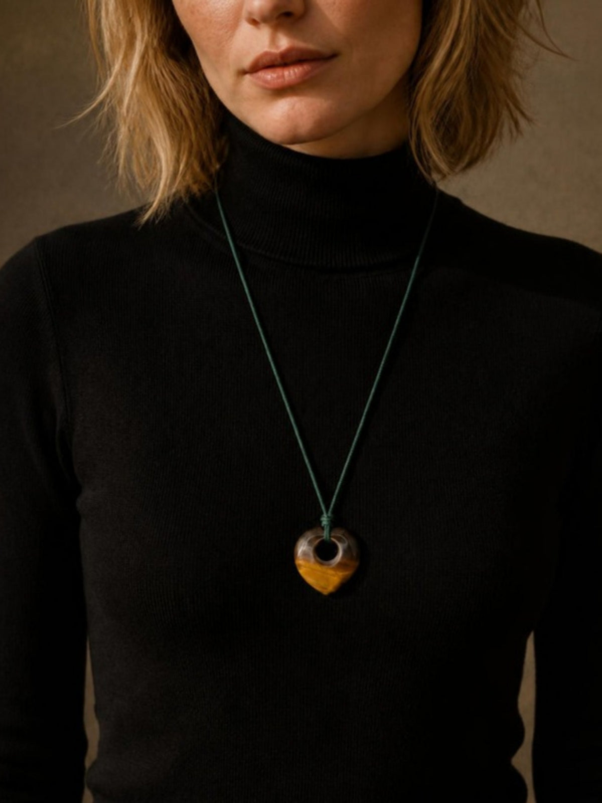 Blonde model wearing the Isla Sol Darkwood Moss Necklace, tiger's eye heart pendant on a dark green cord, over a black turtleneck